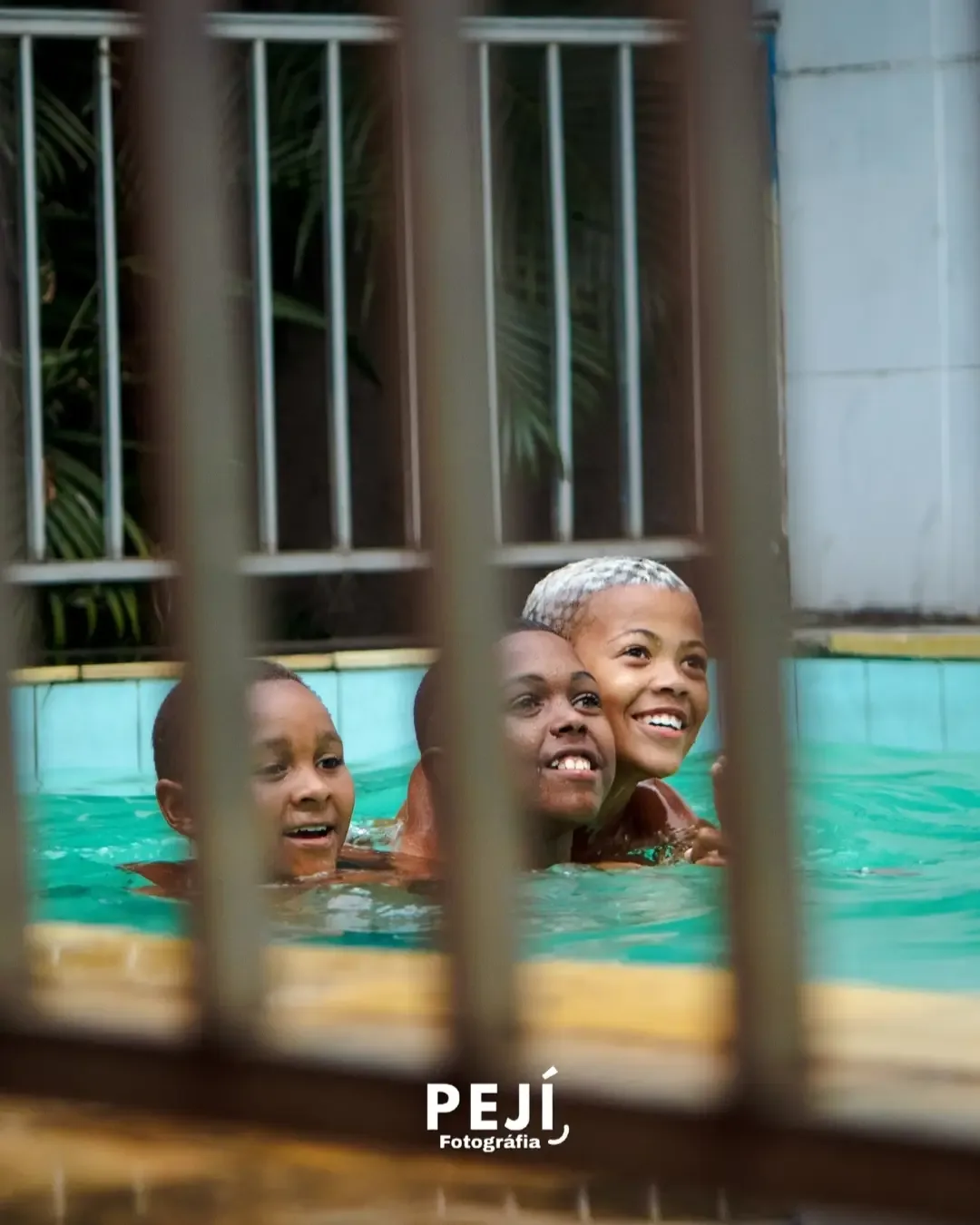 Duas crianças sorrindo na piscina, vistas através de uma grade.