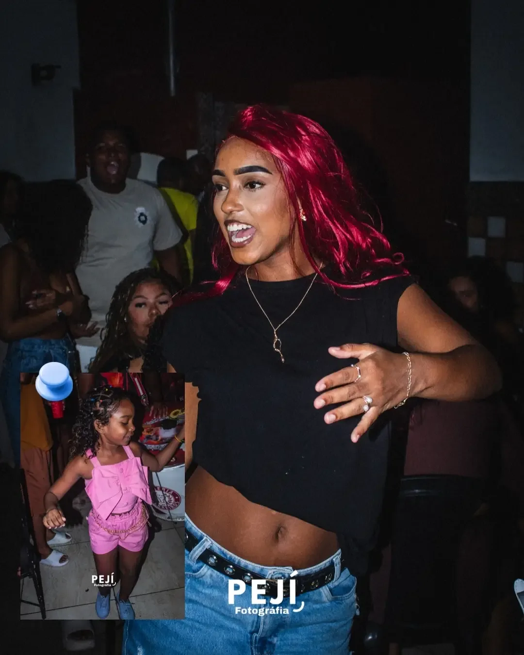 Mulher de cabelo vermelho e blusa preta durante uma festa, com criança ao fundo.