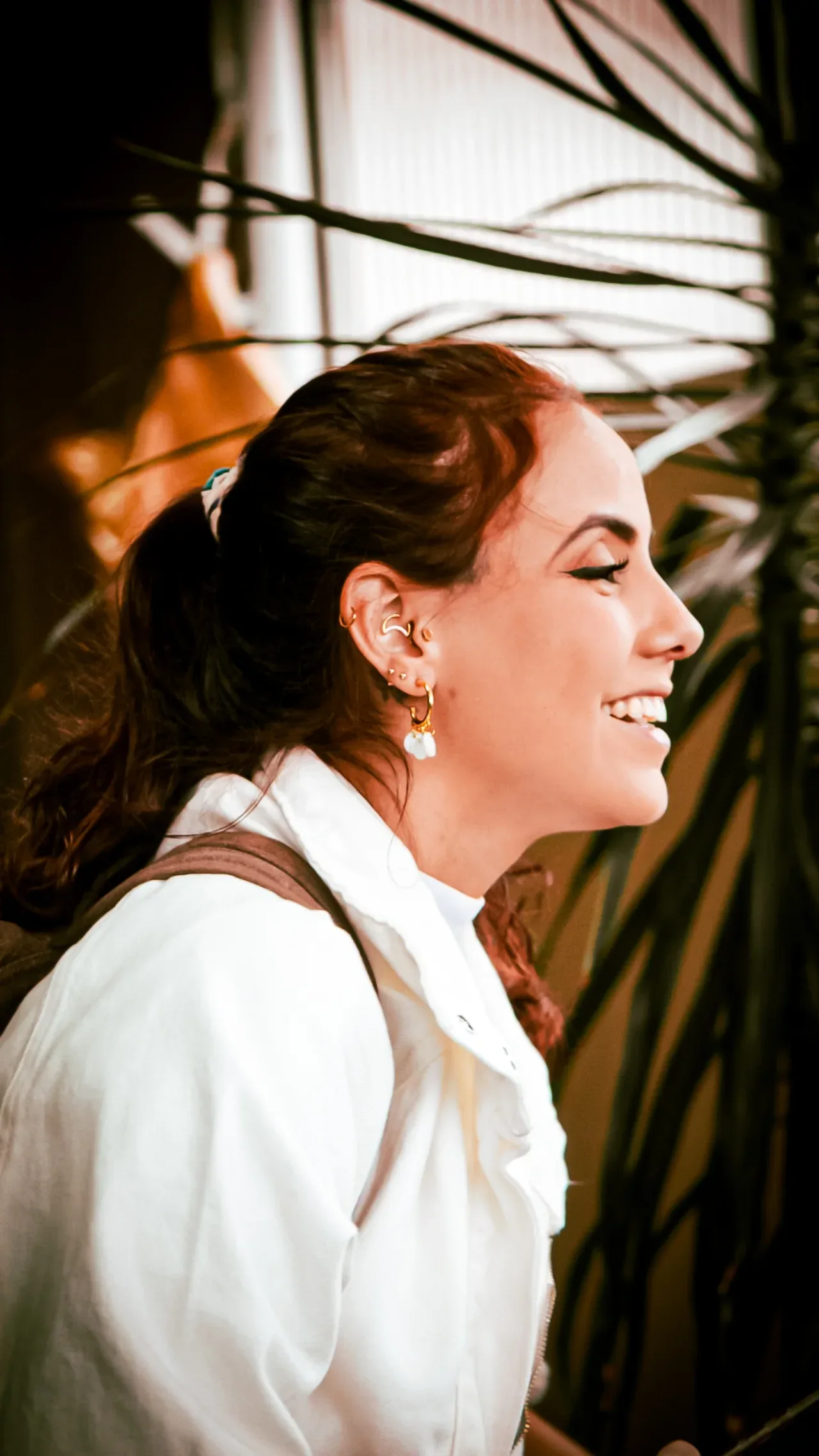 Mulher de roupa branca sorrindo de perfil, iluminada pelo sol junto à vegetação.