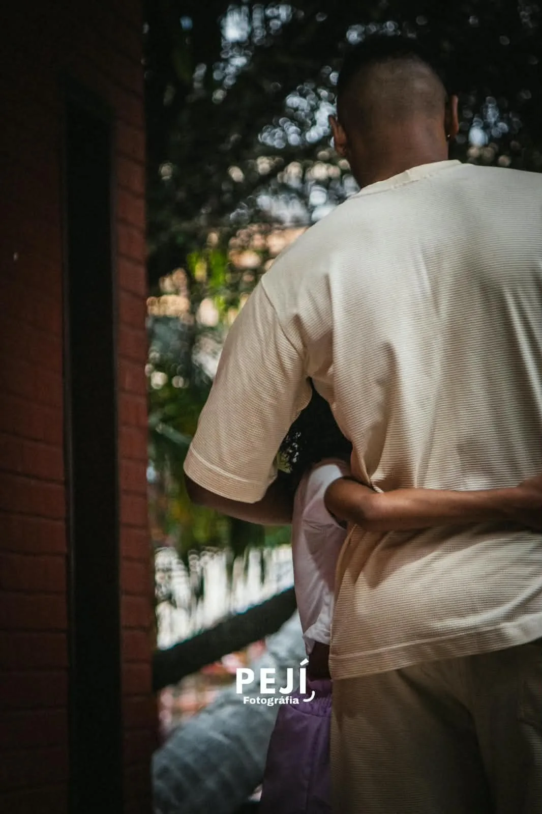 Casal abraçado, visto de costas, próximo a uma porta aberta para o jardim.